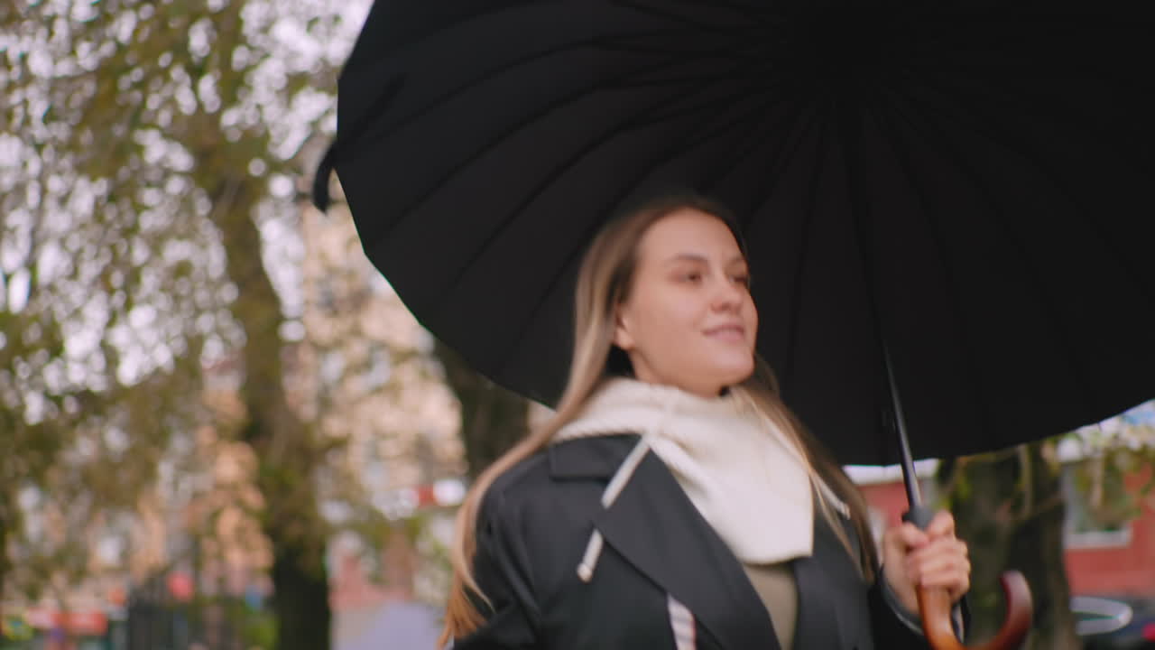 Young woman holding umbrella running during windy day, hair blowing across face, dressed in black coat with white hoodie, walking through city street with autumn trees and blurred buildings in background