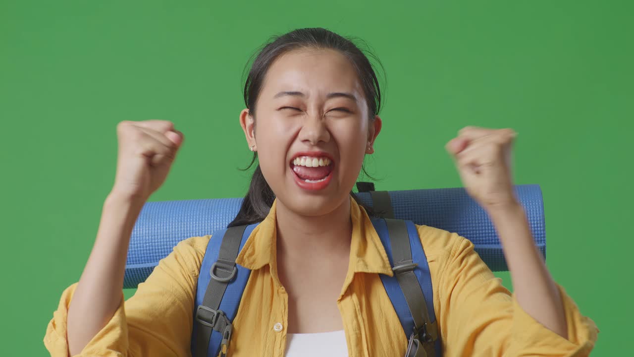 아시아 여성 하이커가 산악 배을 들고 성공을 축하하는 목표를 외치며 스튜디오에서 녹색 화면 배경에 서 있습니다.