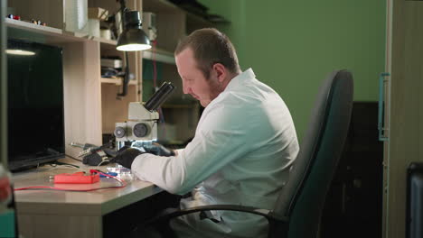 um técnico focado em uma bata de laboratório branca examina um circuito sob um microscópio em um laboratório bem equipado, o espaço de trabalho possui instrumentos científicos, uma lâmpada de mesa e várias ferramentas