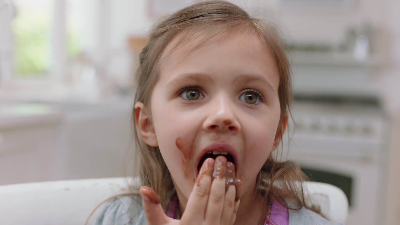 una ragazzina carina con le mani coperte di cioccolato che lecca le dita divertendosi a cuocere in cucina un bambino cattivo che si gode un gustoso dolce a casa