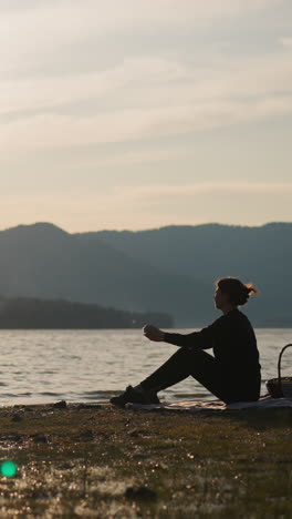 mujer lanza piedra al lago aplaudiendo las manos al atardecer. dama con canasta de picnic disfruta de momentos en la orilla del río con impresionantes vistas de las montañas forestales