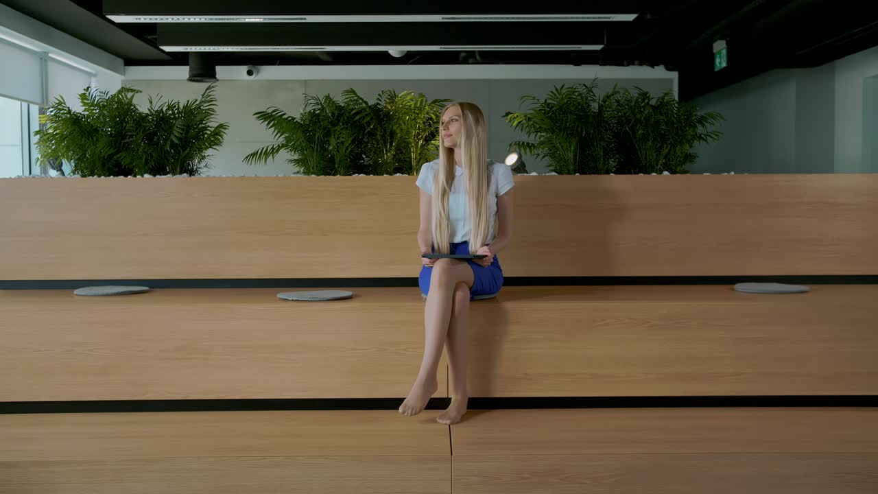 mujer relajada con tableta en la oficina. joven mujer elegante sentada descalza en una tribuna de madera en una oficina moderna usando tableta y descansando