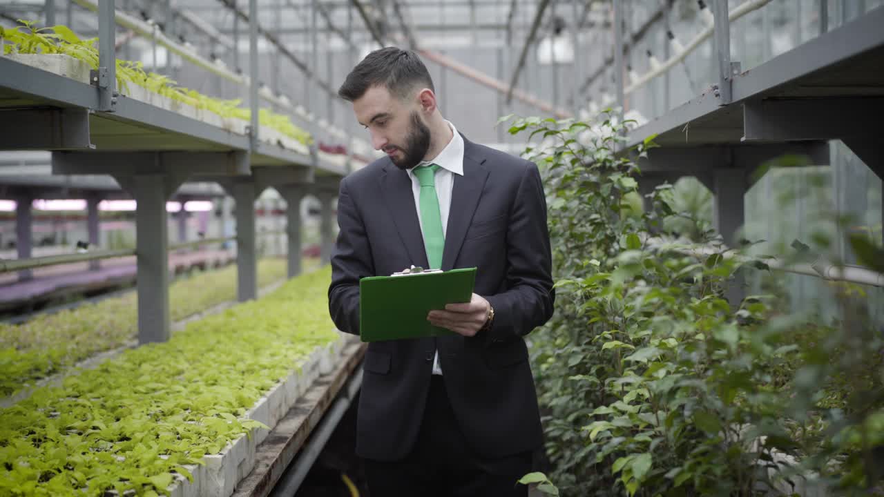 joven hombre caucásico confiado en traje caminando a la cámara en el invernadero y saliendo. hombre de negocios profesional controlando la producción de alimentos vegetarianos orgánicos en el invernador. agricultura, agronomía.