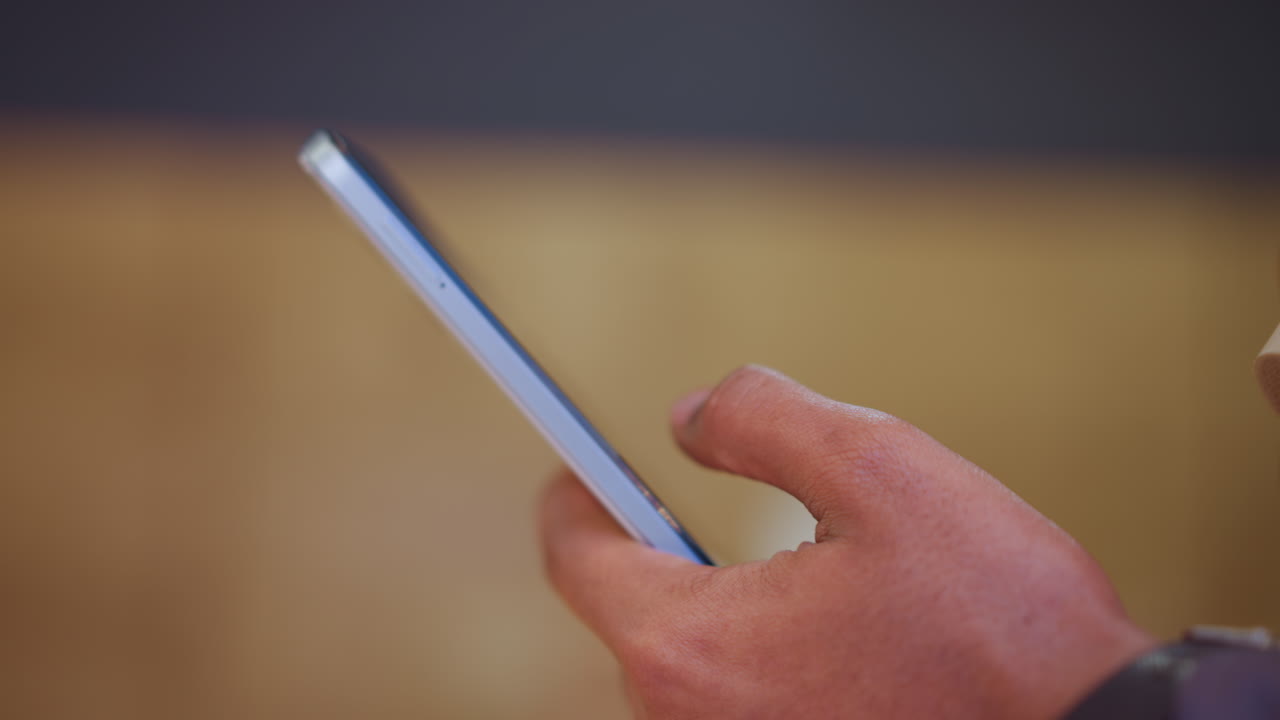 Close-up of male hand holding smartphone with one hand, actively using device in warm ambient setting, sharp focus on fingers and side of phone, suggesting digital interaction, browsing or texting action