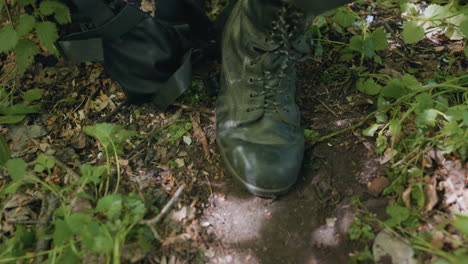 close up view of military man right boot bending at toe while flexing on forest floor beside black bag with scattered leaves and loamy soil under filtered light conveying rugged field operation