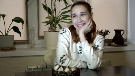 joven sonriente sentada en la mesa cerca del pastel, mirándolo, sonriendo y suspirando. retrato. concepto. 4k.