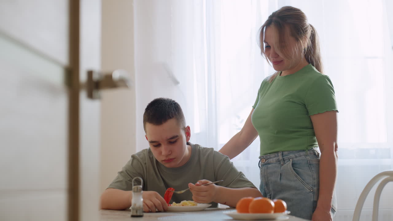 Aunt stands next to teen eating lunch at home, watching in admiration with warm smile, natural daylight through curtain creating tender family moment of care, support, and everyday connection