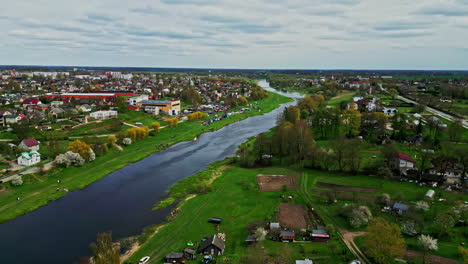 Bauska,-latvia,-showcasing-a-river,-houses,-and-green-fields-on-a-cloudy-day,-aerial-view