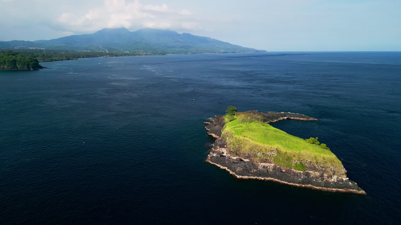 Letecký záběr dronem ukazuje izolovaný ostrov poblíž Bali pokrytý zelenou trávou, obklopený hlubokým modrým oceánem, členitými okraji vulkanických skal a klidnou tropickou atmosférou se vzdálenými horami