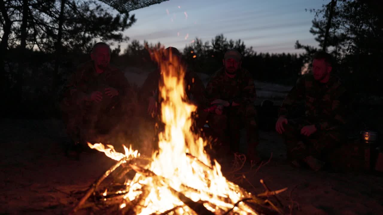 un equipo de hombres con ropa de camuflaje está sentado en una colina en el campo por la noche, calentándose junto a una fogata encendida, fritando salchichas para la cena, una salida masculina, relajándose después del trabajo, el servicio, la caza
