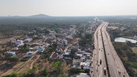 high-angle aerial view of rural development: a major road carrying vehicles, bordered by homes, palm trees, and a pond, with a scenic mountain backdrop