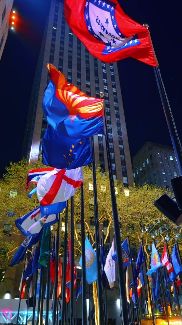 Flags waving outside a skyscraper at night, New York City, Upward low angle perspective of multiple colorful flags fluttering on poles below an illuminated stone highrise.