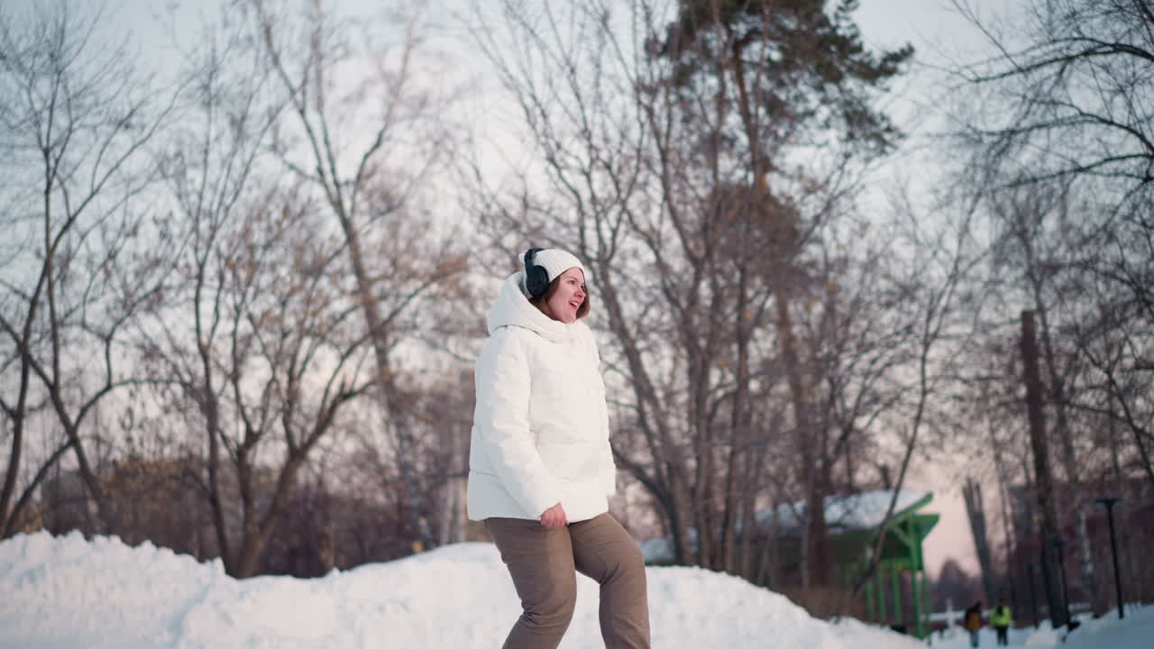 Fair skinned girl dancing on snowy path under twilight sky wearing white puffer coat beanie headphones swaying arms legs in joyful rhythm among bare trees snow mounds conveying cheerful winter vibe