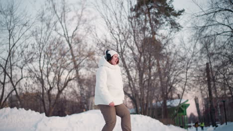 fair skinned girl dancing on snowy path under twilight sky wearing white puffer coat beanie headphones swaying arms legs in joyful rhythm among bare trees snow mounds conveying cheerful winter vibe