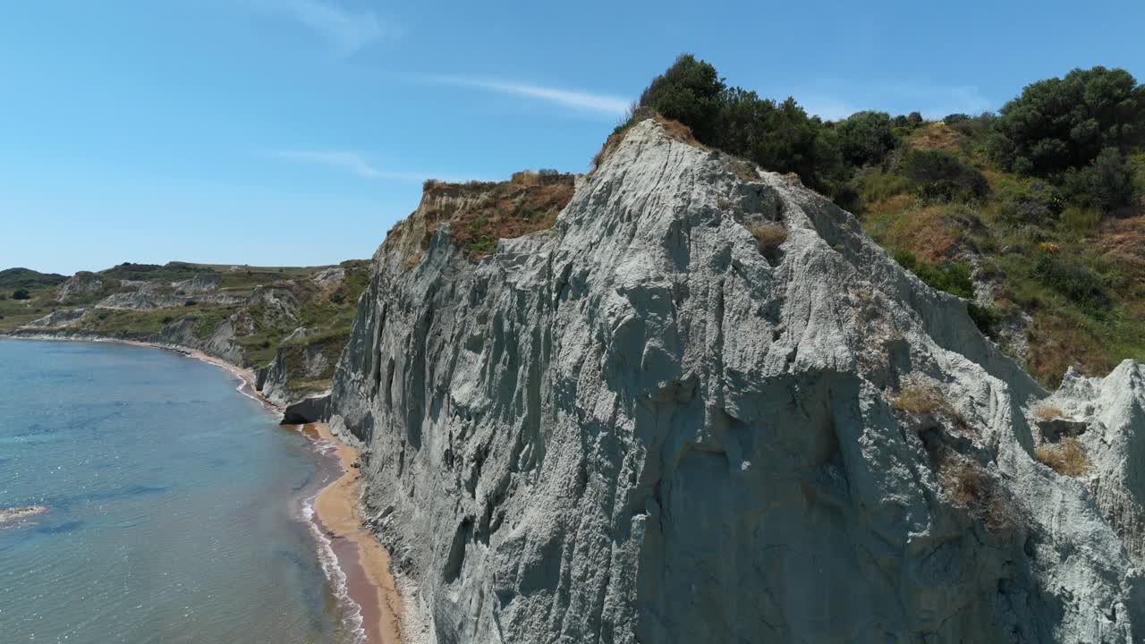 Stunning aerial shot of XI Beach, Kefalonia, featuring unique grey cliffs, vibrant red sand, and sparkling blue waters under bright Mediterranean sunshine.