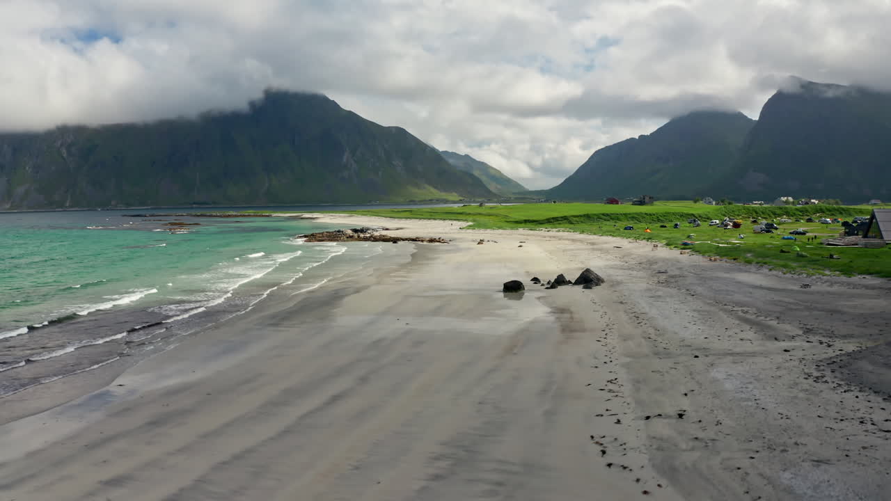 Aerial drone shot over the scenic Nordic Landscape of the Lofoten Islands in Norway. High view of green fields and the long stretching white sanded beach and the turquoise sea.