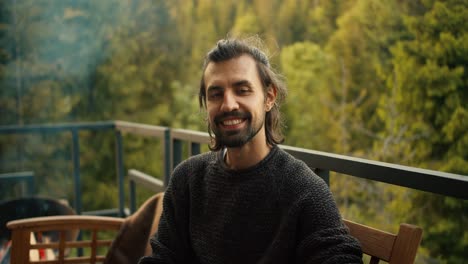 un joven con un suéter verde mira a la cámara y sonríe mientras está sentado en el sofá con el telón de fondo de las montañas y el bosque de coníferas