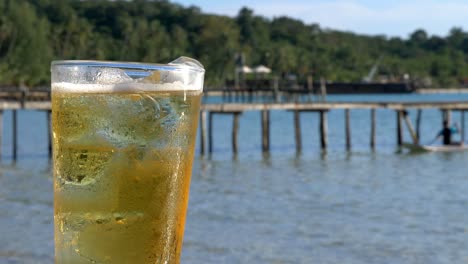 a closeup of a beer, on the beach, slightly angled with a pier in the background cropped