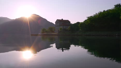 the sun rising from behind a mountain range over the bay of kotor, montenegro, its light reflecting in the water and shining on a mansion and a small coastal town on its shores, aerial fly-above shot