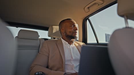 a confident brunette man with black skin and a beard in a light brown shirt holds a laptop in front of him and types there during his business trip in a modern car outside an urban area
