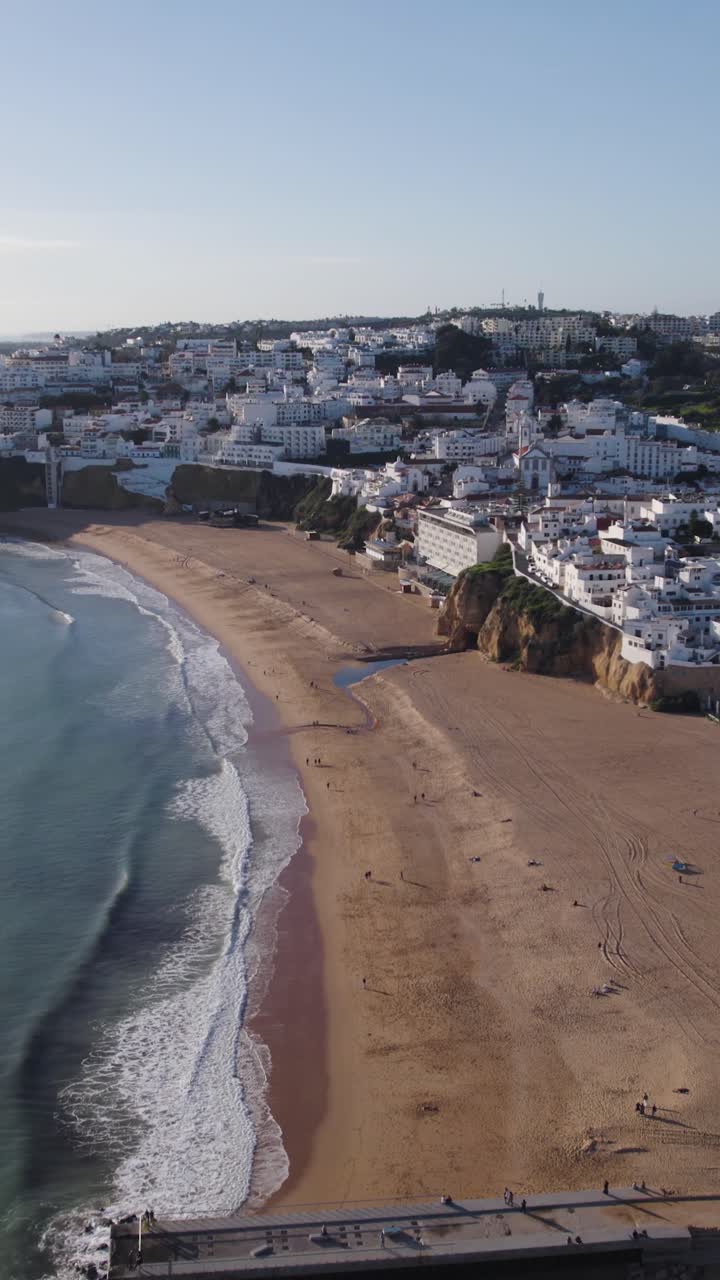 Vertikale Drohnenaufnahme mit Vorwärtsfahrt über einen leeren Strand in Albufeira mit sanften Wellen, die am Ufer brechen.