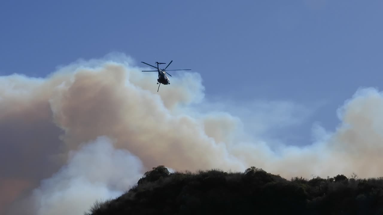 Fire emergency helicopter during the day flying over fire in the Los Angeles hills with smoke in California, USA, handheld shot