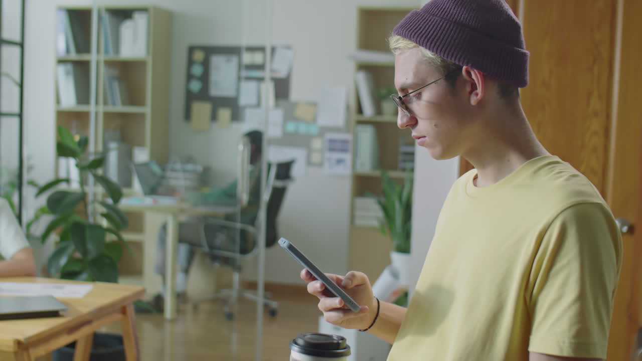 Gen Z Guy Holding Coffee and Scrolling on Phone in Office