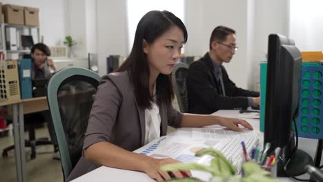 closeup portrait of concentrated asian woman executive typing data while doing paperwork on the computer in the office with other coworkers