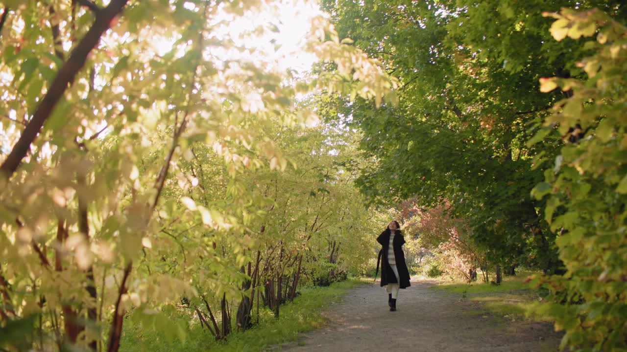 Long shot of autumn lover twirling under sunlit arboretum canopy, coat flowing as she turns along leafy path, golden trees framing joyful spin, soft morning light, serene mood, quiet weekend wander