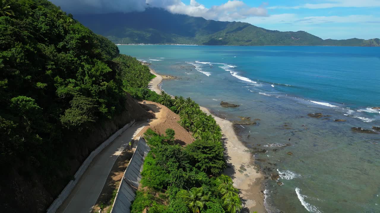 Curving aerial shot in Matawe, Dingalan, Aurora, revealing a hillside road draped in the shifting shadows of tall trees, framed by lush green hills and the coastal landscape
