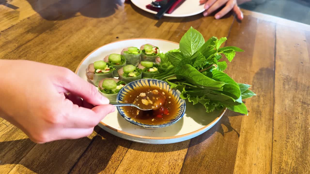 A hand mixes Vietnamese dipping sauce with fresh herbs and vegetables on a wooden table in a Bangkok restaurant