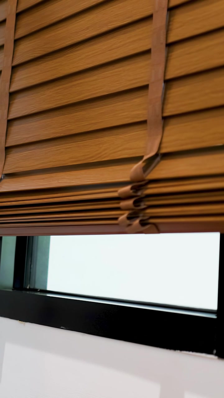 Wooden venetian blinds slowly rise upward against a white wall, revealing a bright window view
