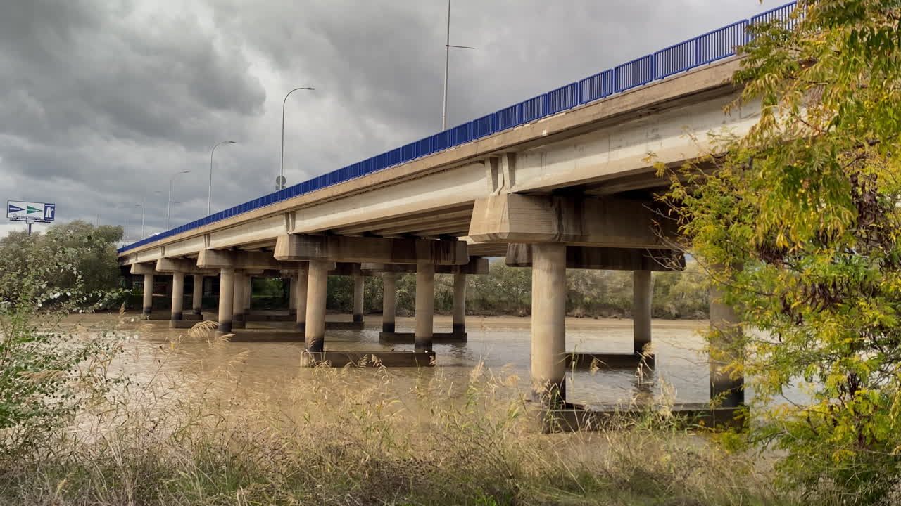 Joen virtaus on hidasta ja ruskeaa sillan alla, ja betoniset pilarit katoavat etäisyyteen luonnon ja infrastruktuurin hiljaisessa tasapainossa.