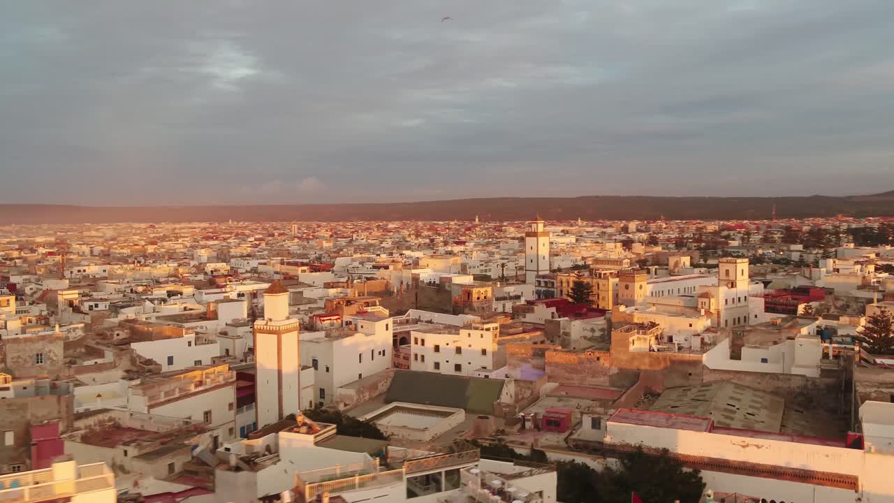 Aerial drone shot of Essaouira's white cityscape and mosque minaret bathed in warm golden sunlight. Establishing shot of Moroccan urban architecture and rooftops