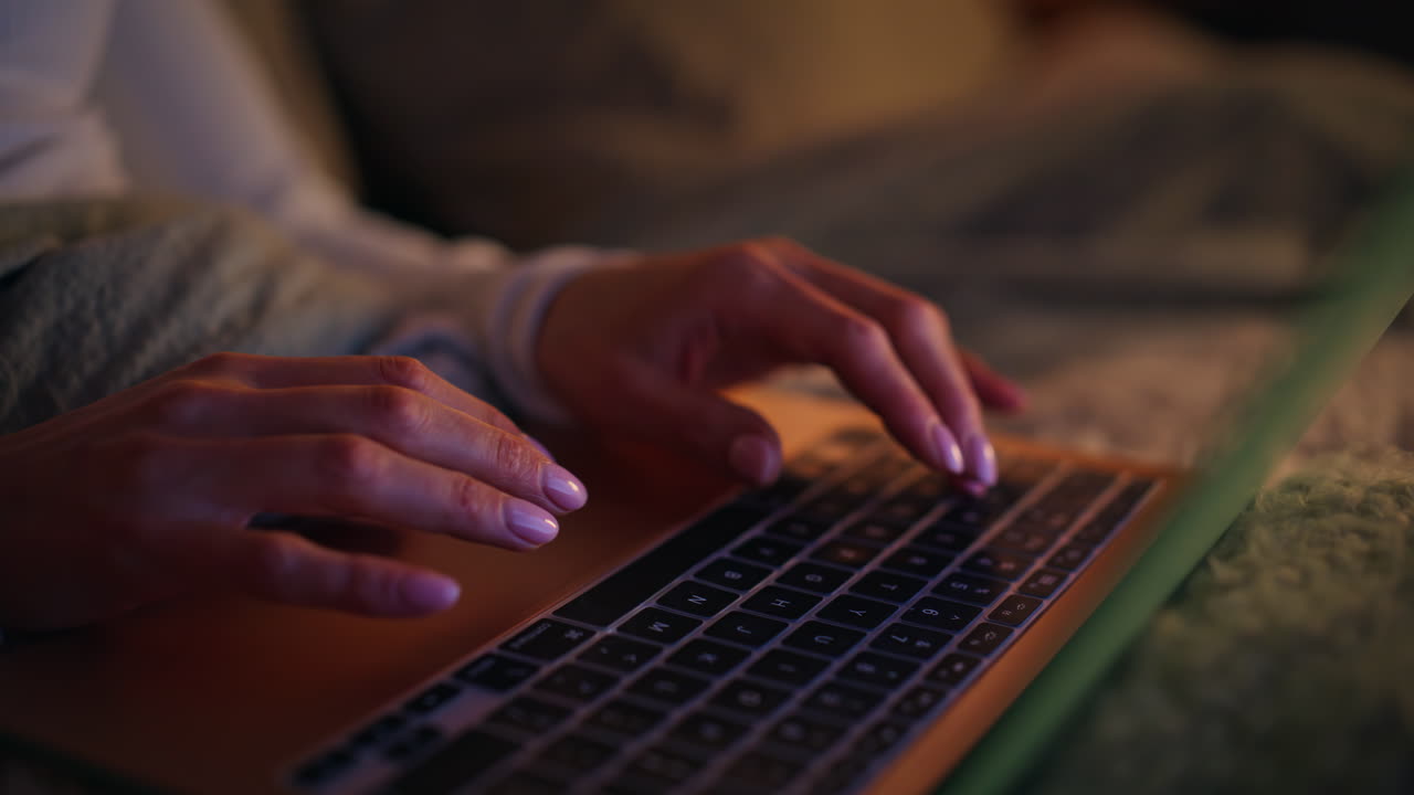 Nærbillede af kvinde, der arbejder på bærbar computer sent om aftenen liggende i seng derhjemme. Kvindens hænder taster.