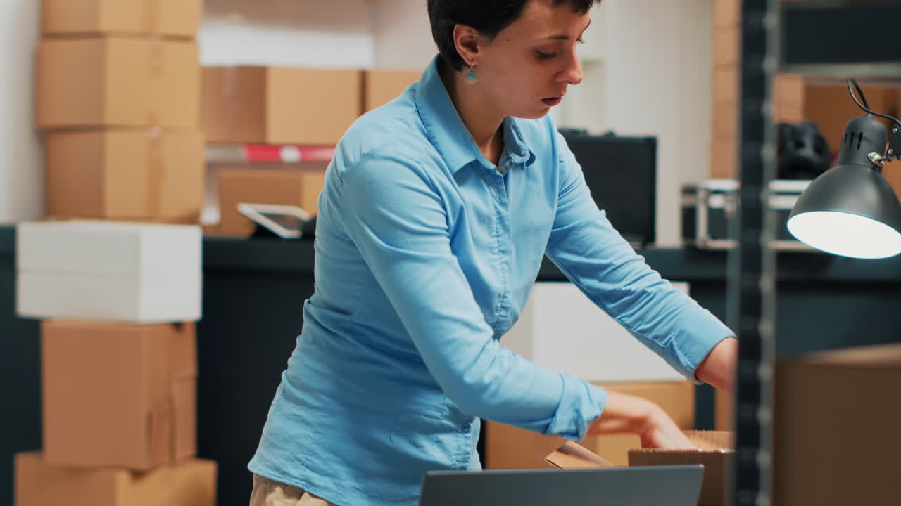 Woman working on laptop in warehouse with inventory and boxes