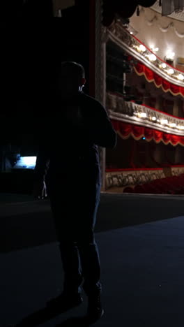 Theater actor has rehearsal before performance. Dark light behind scene. Man in formal clothes with handfree microphone. Classic theater concept. Vertical video