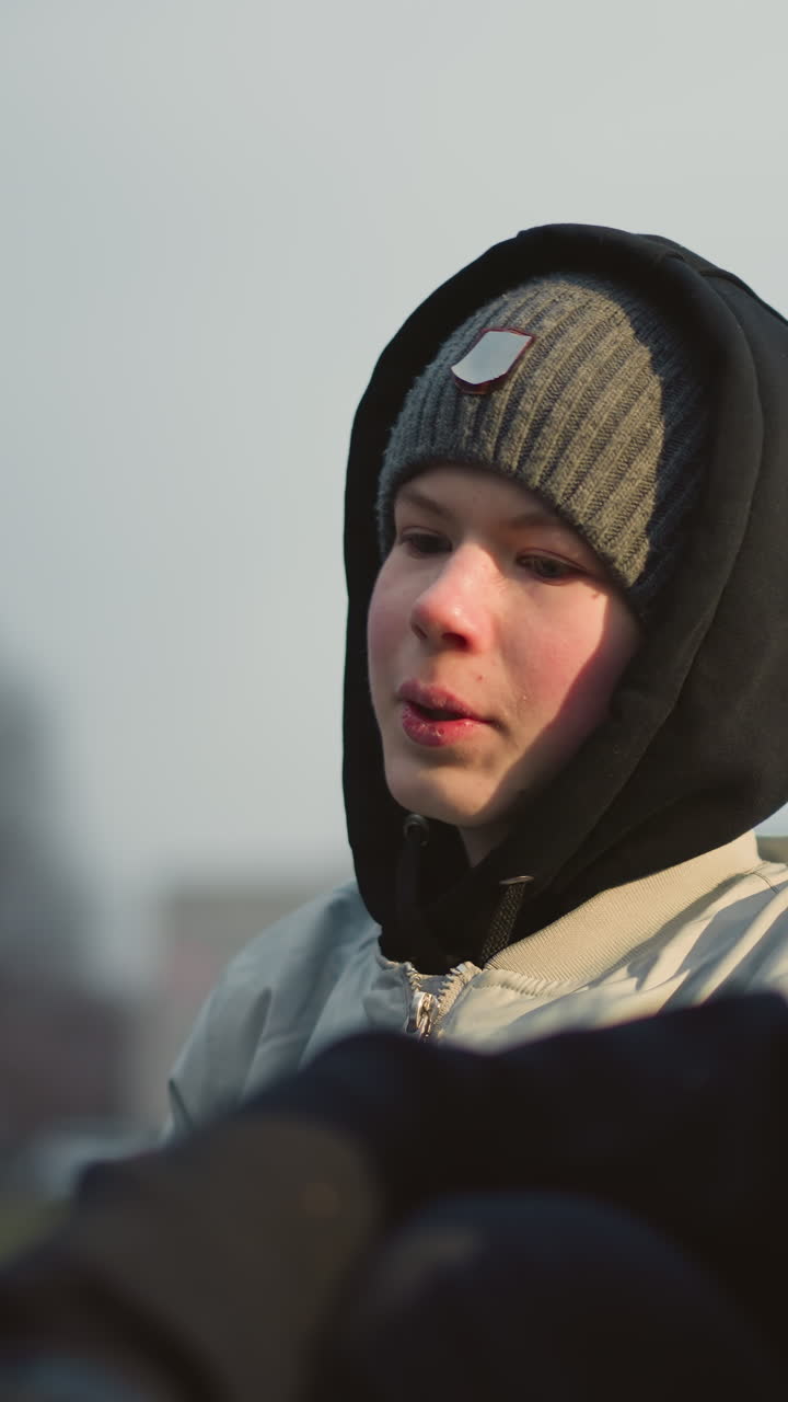 Close-up of a young boy in a gray beanie and black hoodie, intently talking to another person in black, with a blur background