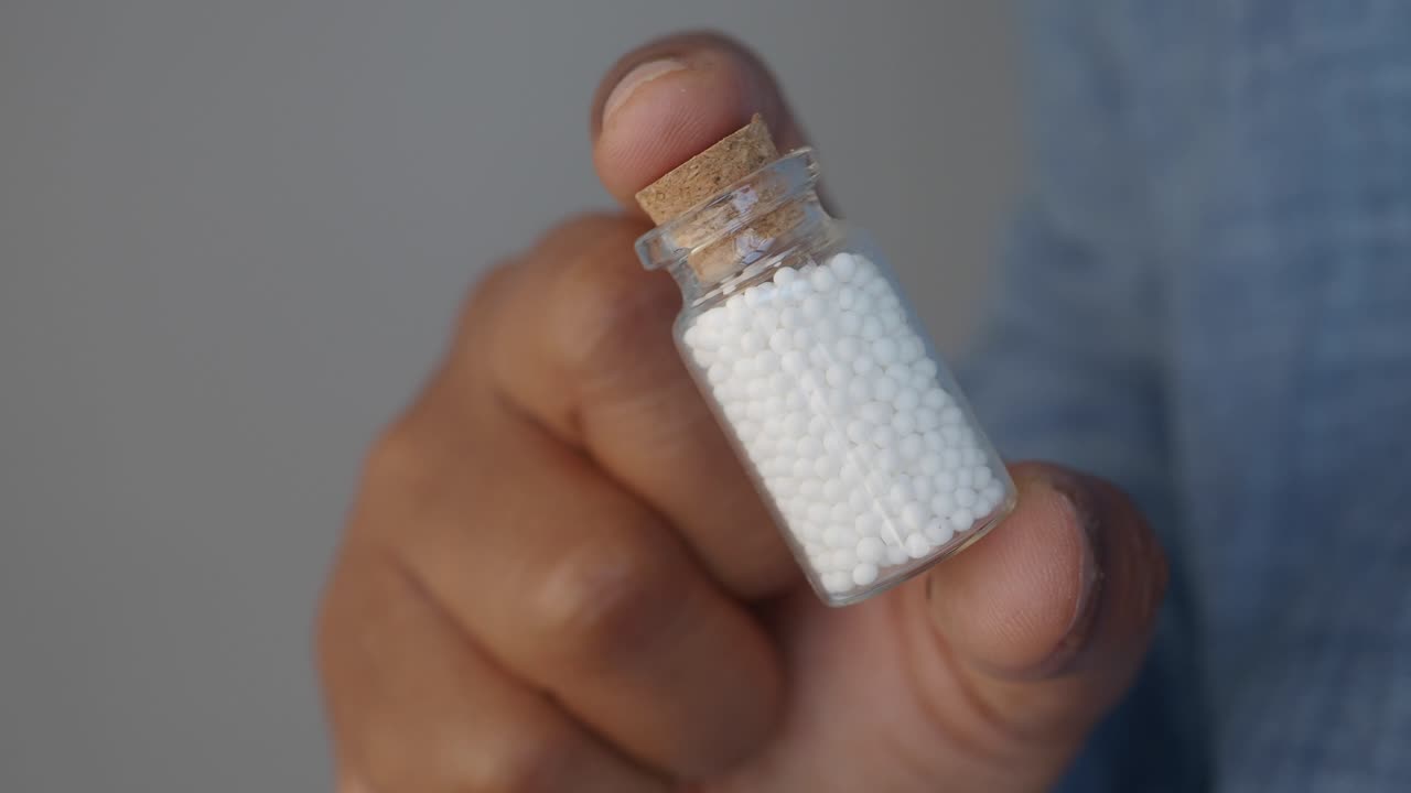 primer plano de una mano sosteniendo una pequeña botella de vidrio llena de pastillas blancas