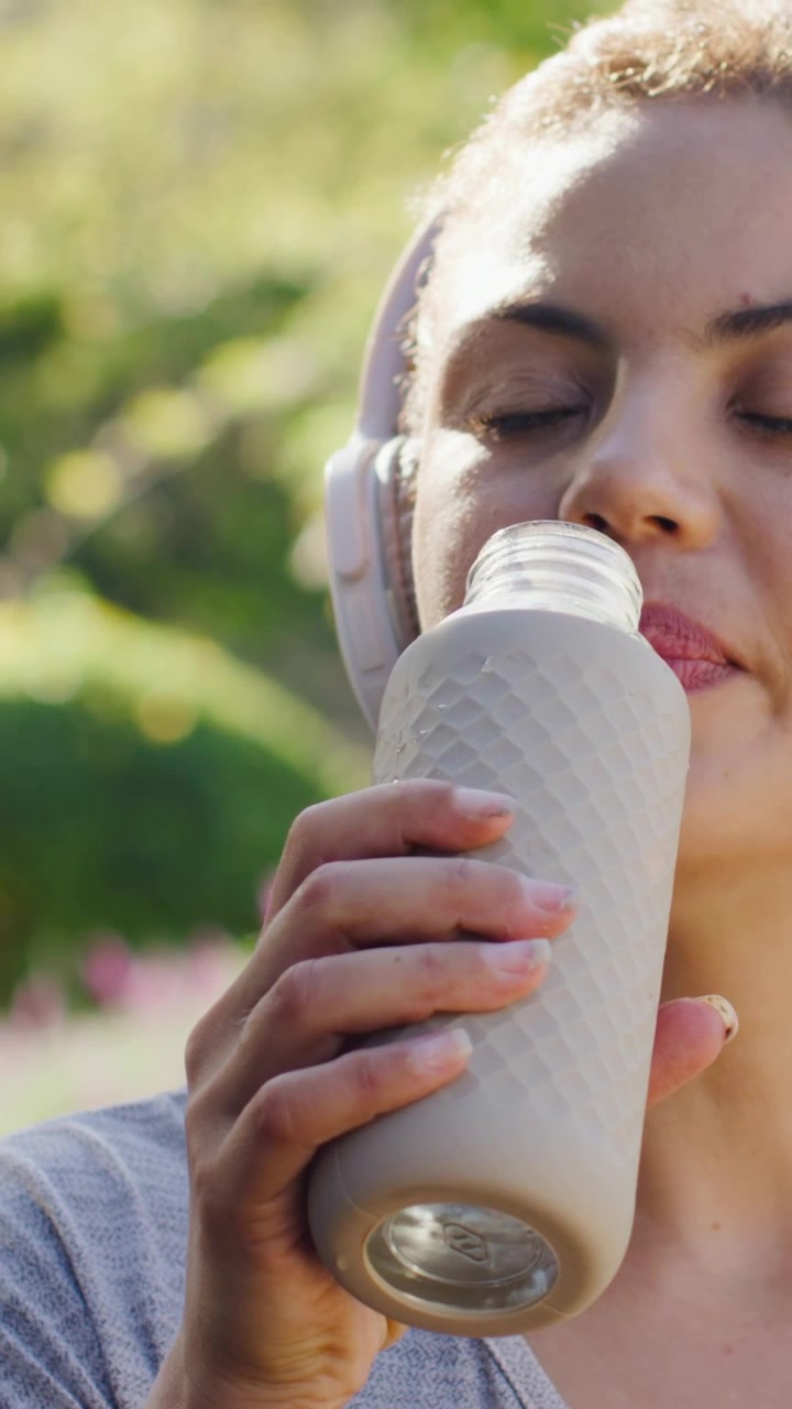 Video verticale: Donna con cuffie che sorseggia al sole da una bottiglia beige testurizzata in giardino