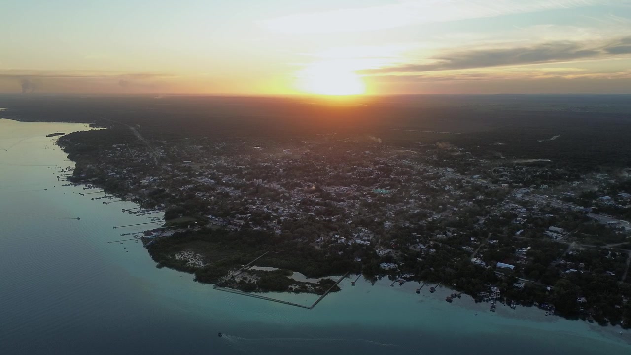 Tomada de drone descendo em direção à costa da cidade de Bacalar ao pôr do sol na hora dourada, revelando a lagoa e a vila à beira do lago em Quintana Roo, México.