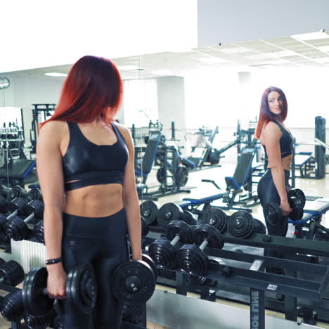 Beautiful fitness woman with dumbbells. Smiling female athlete looks on her sporty figure in the mirror on sports center background. Pretty muscular girl near rows of dumbbells.