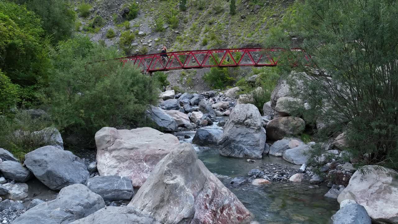 A mountain river flowing over rocky terrain in Theth, Albania, aerial dolly up stream as person walks across Thethi bridge