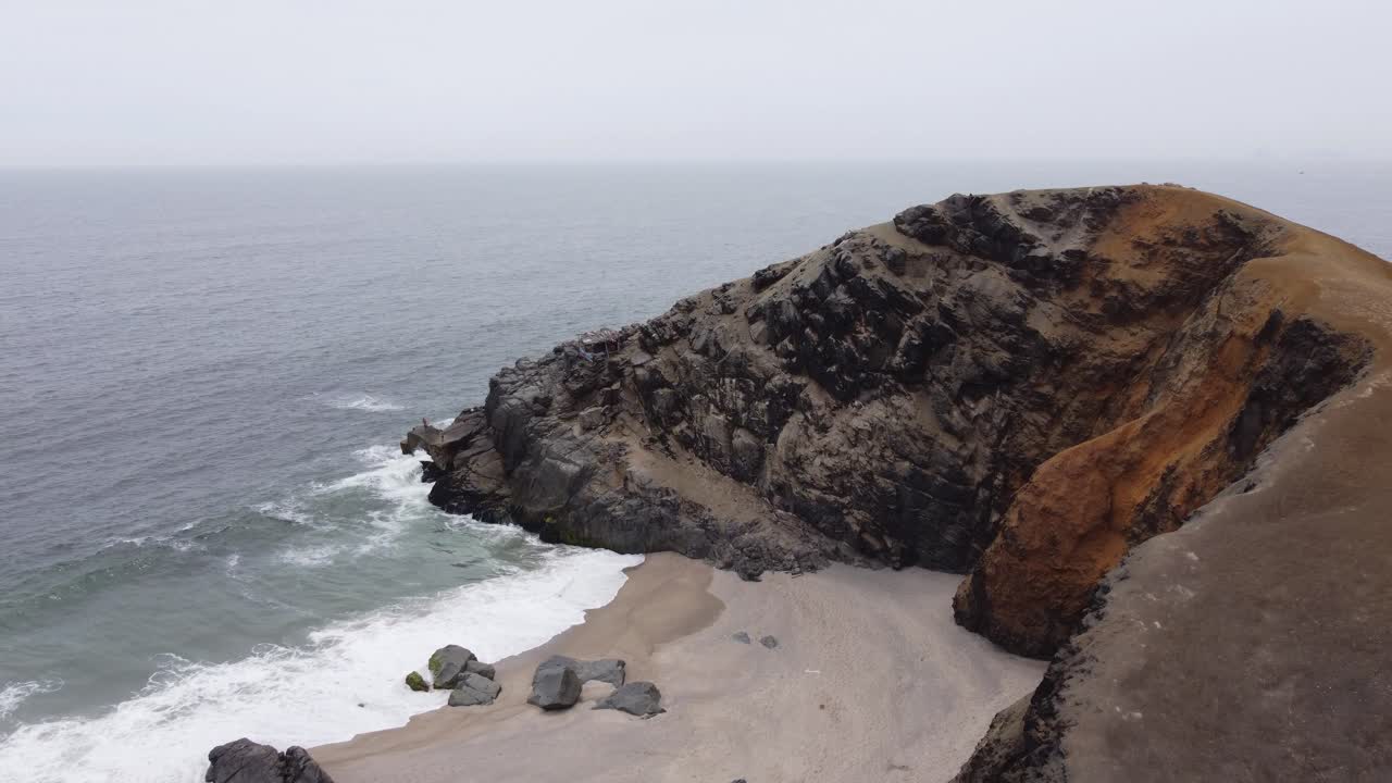 A large rocky outcrop on the edge of the ocean. Drone slowly flies towards the cliff with the sea horizon behind it. Located in Punta Negra beach in Lima, Peru.
