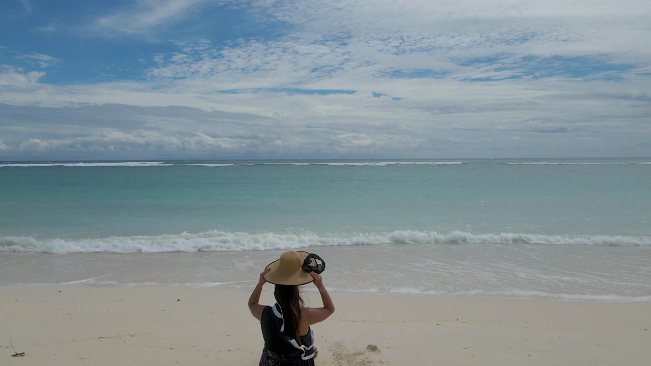 Uma mulher relaxa numa praia tropical intocada em Gili, Índia, colocando um chapéu de palha enquanto observa as ondas turquesa do oceano. Perfeito para conteúdo de viagens e bem-estar