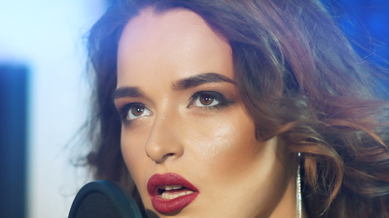 Face of an attractive singer with wavy hair and long earrings close to the microphone. Pretty woman singing in the recording studio. Close-up