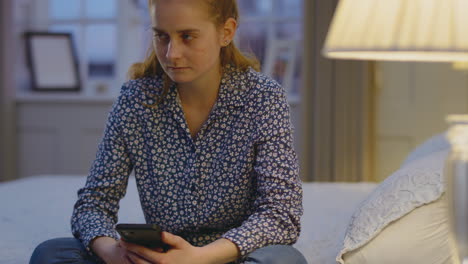 Mujer-Joven-Deprimida-En-Casa-Sentada-En-El-Dormitorio-Por-La-Noche-Con-Un-Teléfono-Móvil-Con-Aspecto-Triste,-Filmada-En-Cámara-Lenta