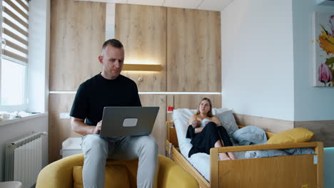man having problems working at laptop. he shows the gadget to a woman sitting at backdrop feeding a baby.