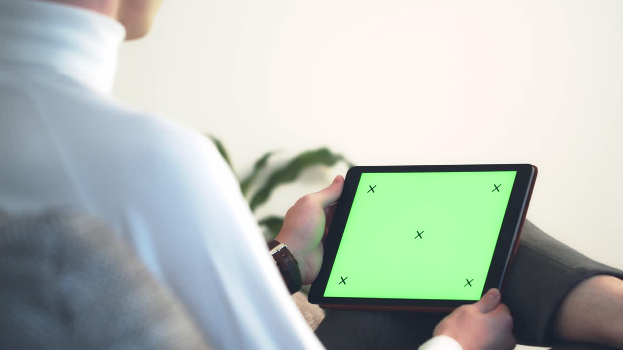 흰색 셔츠를 입은 남자가 아늑한 소파에 앉아 검은색 태블릿 장치의 크로마 키를 손에 들고 녹색 화면을 스크롤합니다.