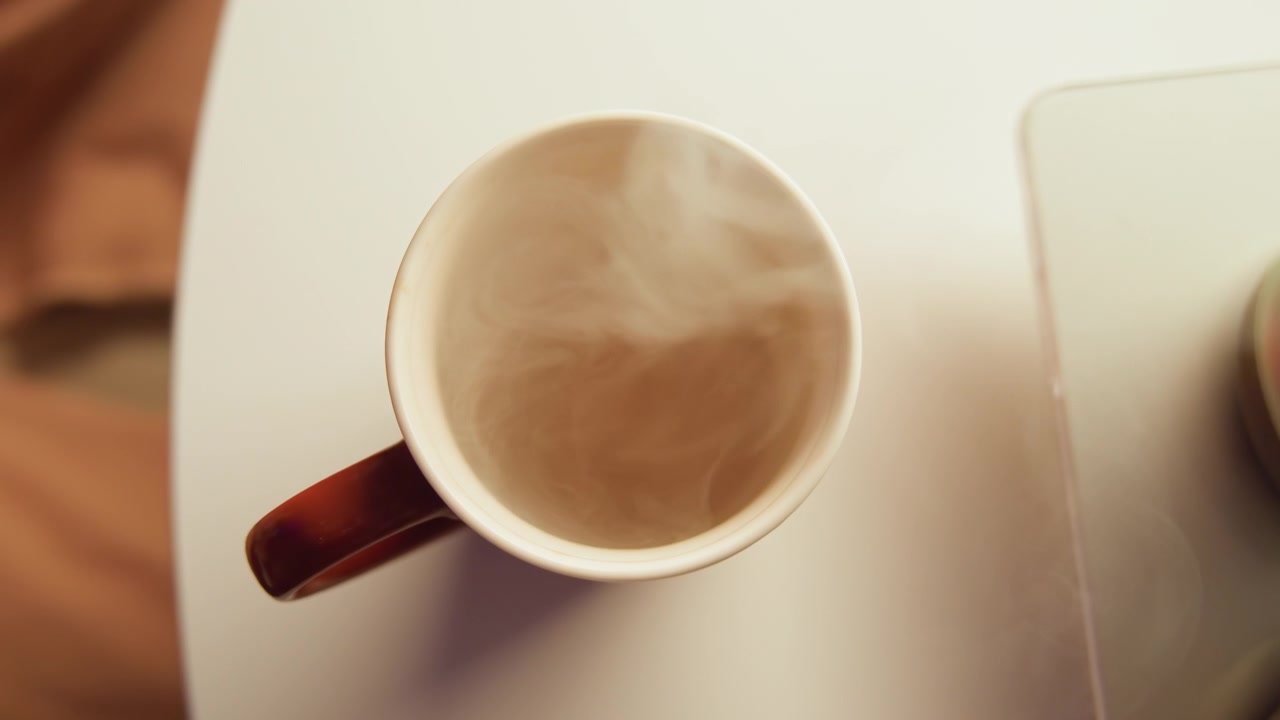 Una vista dall'alto mostra una mano che afferra una tazza di caffè in ceramica per il manico accanto all'angolo di un laptop su una scrivania chiara, con vapore che sale, superficie vorticosa, leggera inclinazione e un'atmosfera calma da spazio di lavoro mattutino.